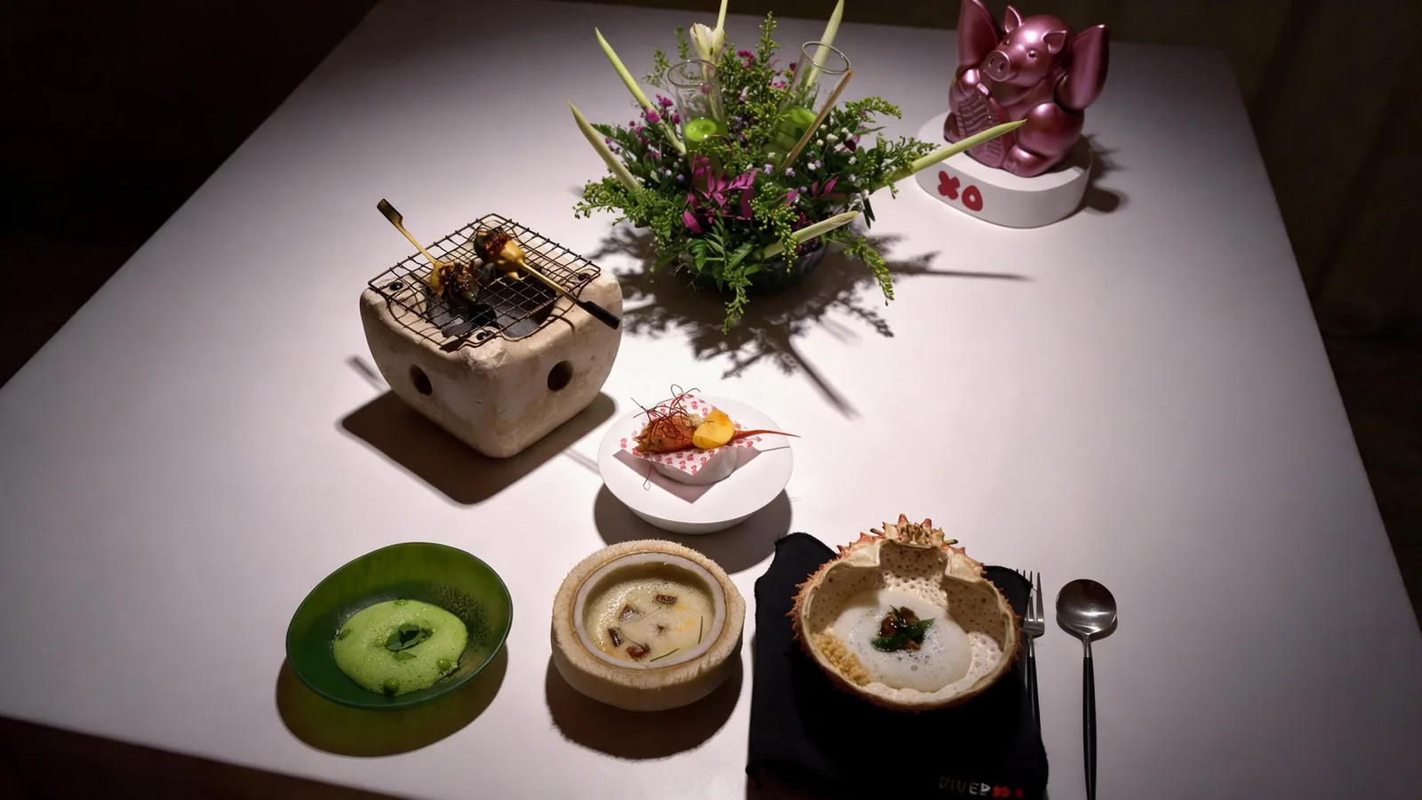 A white table at DiverXO set with a stone grill, a prawn, two soups, a crab shell of foam, a flower centrepiece and a pink pig figurine.
