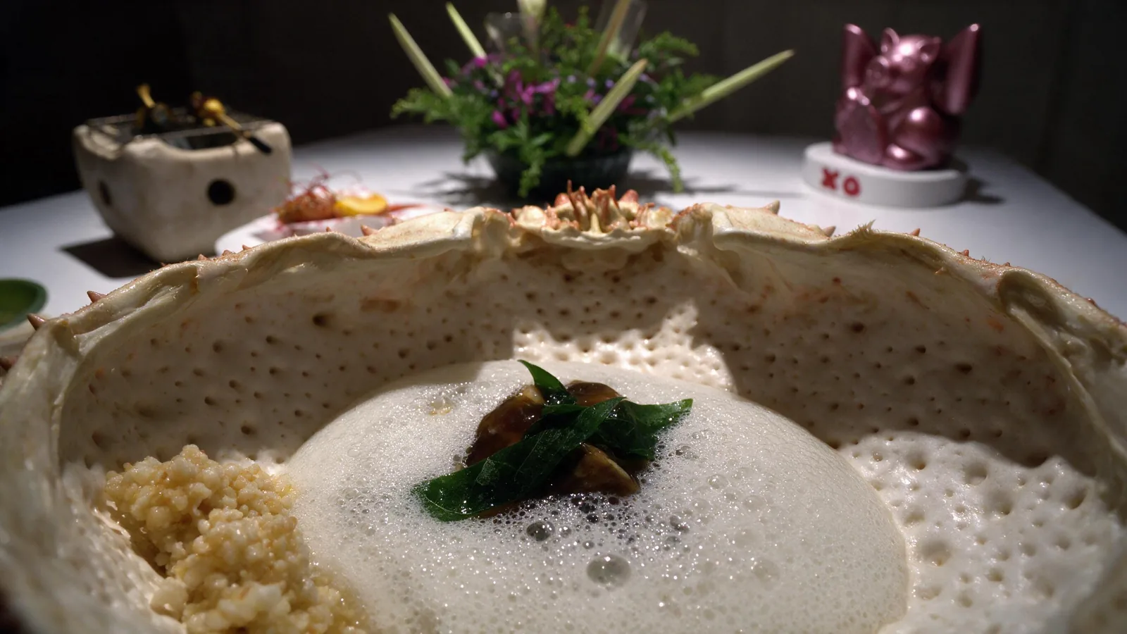Close up inside a crab shell filled with white foam, a dark glazed morsel and a green leaf at the centre.
