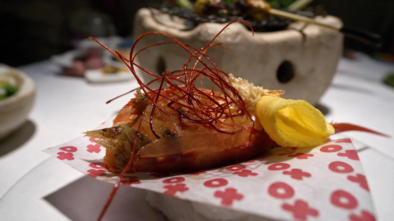 Close up of a grilled prawn under fine red threads beside a yellow crisp, on red and white XO paper.