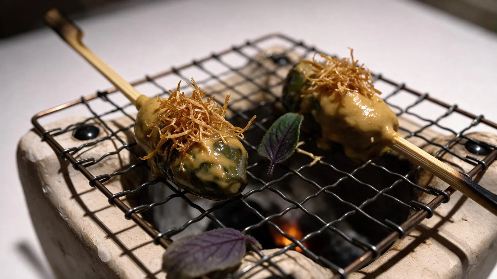 Close up of two skewers coated in ochre sauce and fried threads, resting on a wire grill over glowing charcoal.